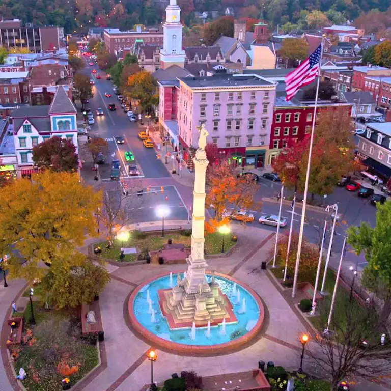 Aerial view of a town square with a fountain monument, American flag, and surrounding buildings at dusk.